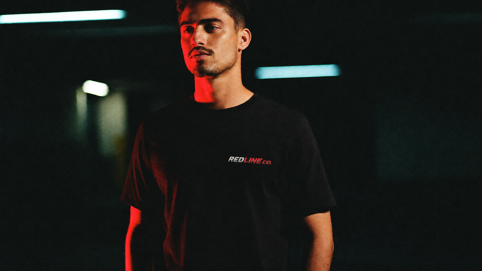 Homem usando camiseta preta com a logo RedLine estampada no peito, em garagem escura sob luz vermelha dramática