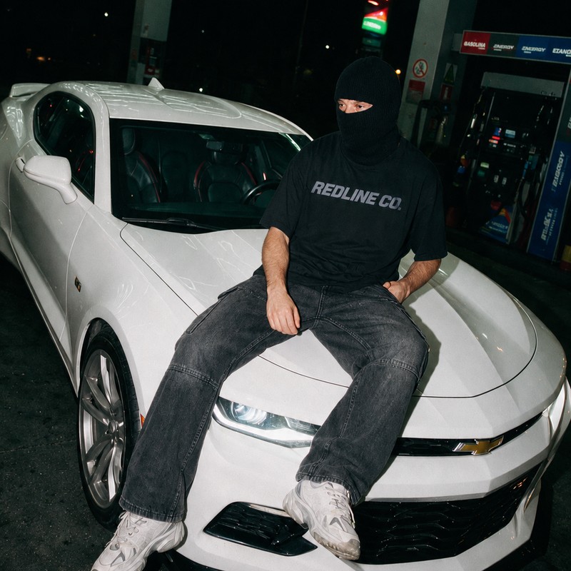 Integrante mascarado sentado no capô do Camaro branco, num posto de gasolina à noite