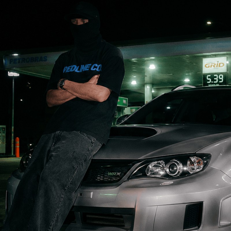 Integrante mascarado de camiseta RedLine, encostado no capô do WRX prateado num posto à noite